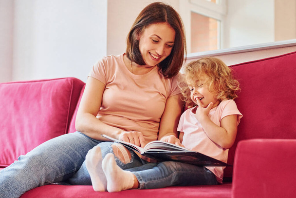 Reading big book. Young mother with her little daughter in casual clothes together indoors at home. - Photo, Image