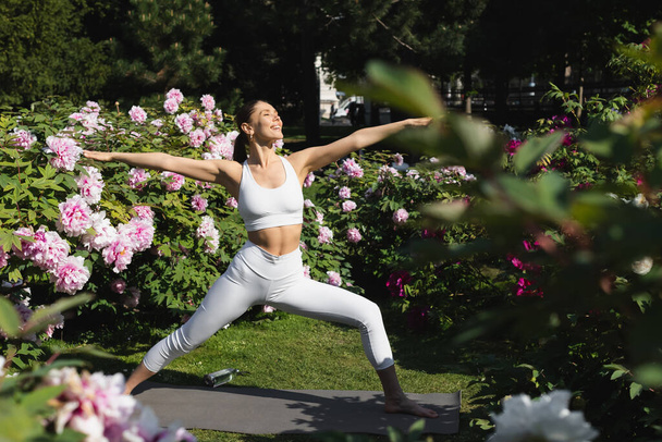 happy sportive woman practicing warrior pose with outstretched hands in park  - Foto, Imagem
