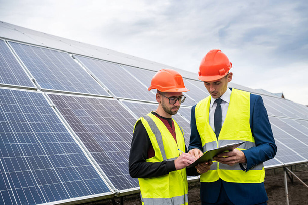 kaksi työvaatteiden ammattilaista keskustelevat aurinkopaneeleista ja niiden vaikutuksista. Vihreän sähkön käsite ja sen vaikutukset - Valokuva, kuva