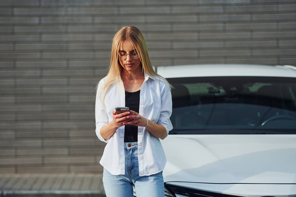 Beautiful blonde in casual clothes with phone near white car is outdoors at sunny daytime. - Photo, image