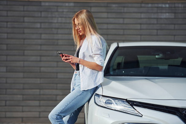 Schöne Blondine in lässiger Kleidung mit Handy in der Nähe weißes Auto ist draußen bei sonnigem Tag. - Foto, Bild