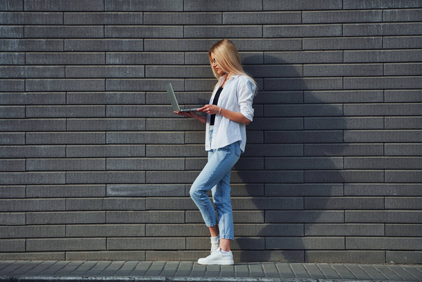 Using laptop. Beautiful blonde in casual clothes is outdoors at sunny daytime. - Photo, Image