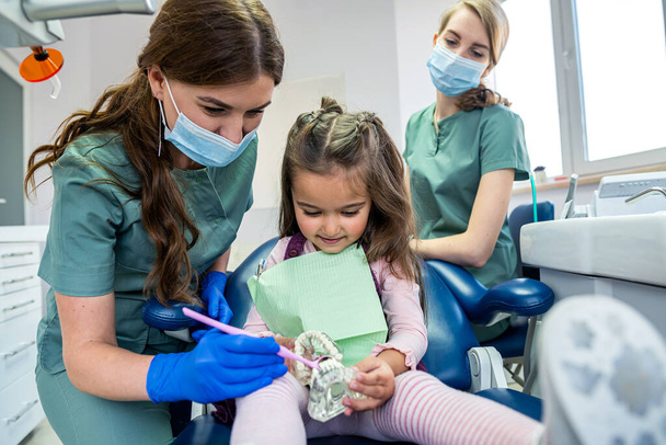 Kinderzahnarzt zeigt am künstlichen Kiefer, wie man Zähne putzt. Kleine Patientin sitzt auf Stuhl und hört auf den Rat eines Zahnarztes - Foto, Bild