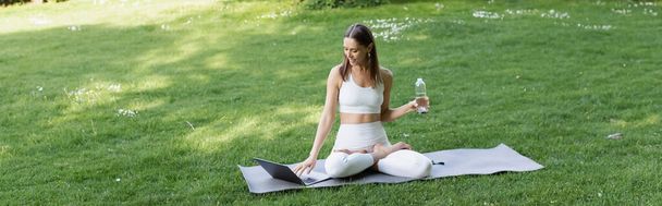 nő fehér sportruházat segítségével laptop, miközben ül lótusz pózol sport palack, banner - Fotó, kép