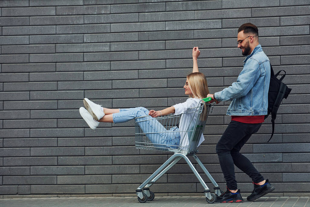 Having fun and riding shopping cart. Young stylish man with woman in casual clothes outdoors together. Conception of friendship or relationships. - Valokuva, kuva