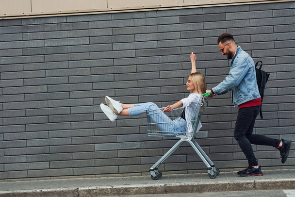 Having fun and riding shopping cart. Young stylish man with woman in casual clothes outdoors together. Conception of friendship or relationships. - 写真・画像