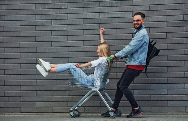 Having fun and riding shopping cart. Young stylish man with woman in casual clothes outdoors together. Conception of friendship or relationships. - Foto, Imagem