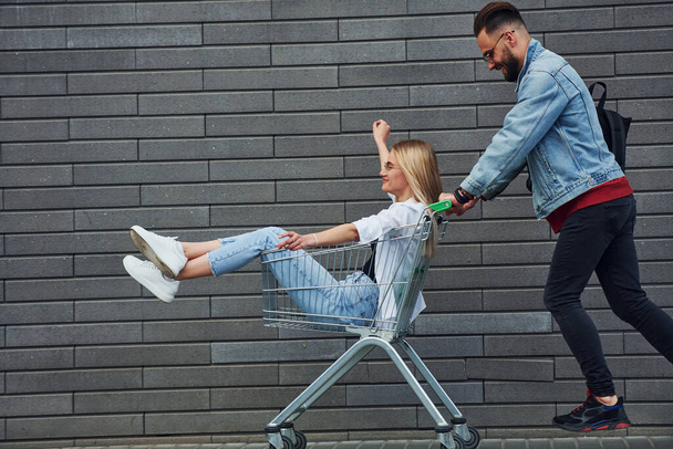 Having fun and riding shopping cart. Young stylish man with woman in casual clothes outdoors together. Conception of friendship or relationships. - Fotoğraf, Görsel