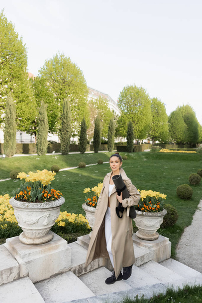 sportswoman in trench coat holding fitness mat while walking on stairs near park vases with flowers - Foto, Imagem