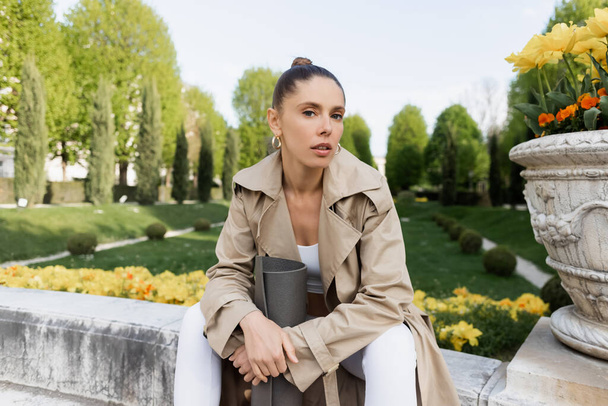 woman in trench coat looking at camera while sitting with fitness mat near park vase - 写真・画像