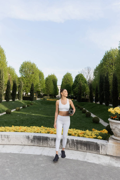 volledige lengte van de vrouw in witte sportkleding houden fitness mat en weg te kijken in park  - Foto, afbeelding
