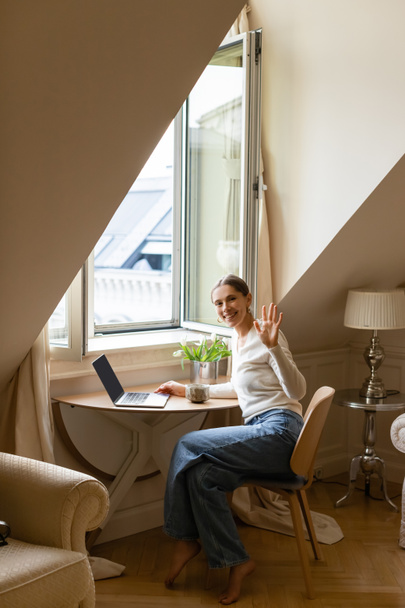 mujer feliz agitando la mano mientras está sentado con el ordenador portátil y taza de arcilla cerca de la ventana - Foto, Imagen