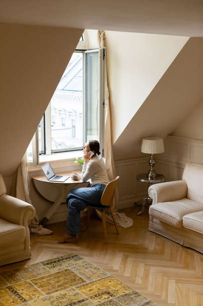 barefoot woman talking on mobile phone near laptop and window in cozy attic room - Photo, Image
