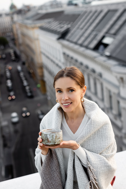 woman covered with shawl holding clay cup and smiling at camera near rooftops on blurred background - 写真・画像