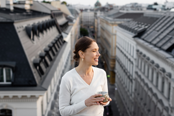 smiling woman with clay cup looking at blurred urban rooftops - Photo, Image