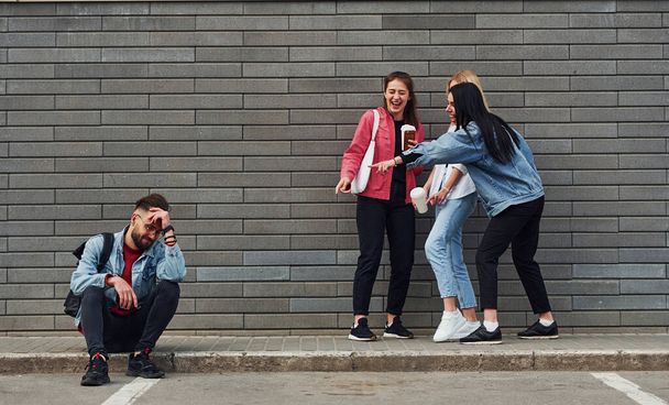 Three women standing and laughing at guy that sitting outdoors near building at daytime. - 写真・画像