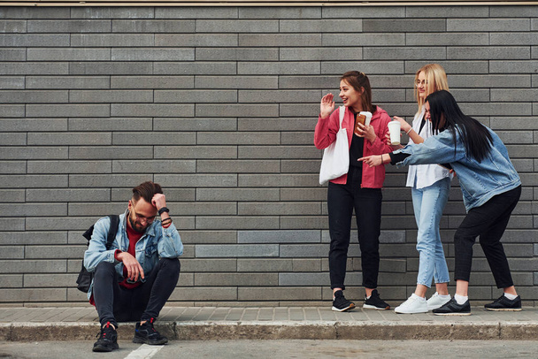 Three women standing and laughing at guy that sitting outdoors near building at daytime. - Valokuva, kuva