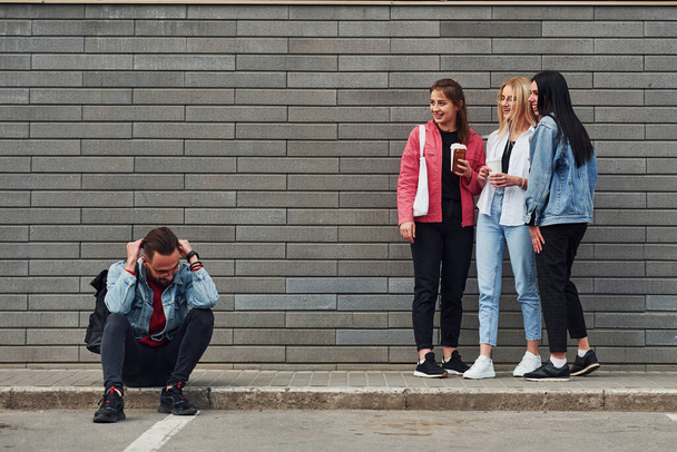 Три женщины стоят и смеются над парнем, который сидит на улице возле здания в дневное время. - Фото, изображение
