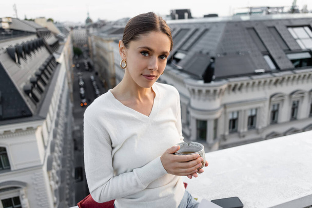 woman in white jumper holding clay cup and looking at camera on rooftop of city building - 写真・画像