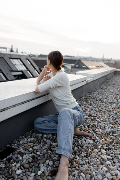 barefoot woman in jeans sitting on rooftop and looking away - Foto, afbeelding