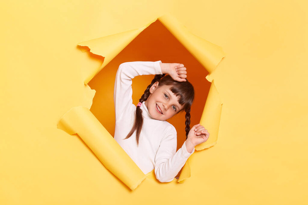 Portrait of funny positive little girl with braids wearing casual shirt looking through torn hole in yellow paper, keeps hands raised, having fun, expressing happiness. - Foto, Bild