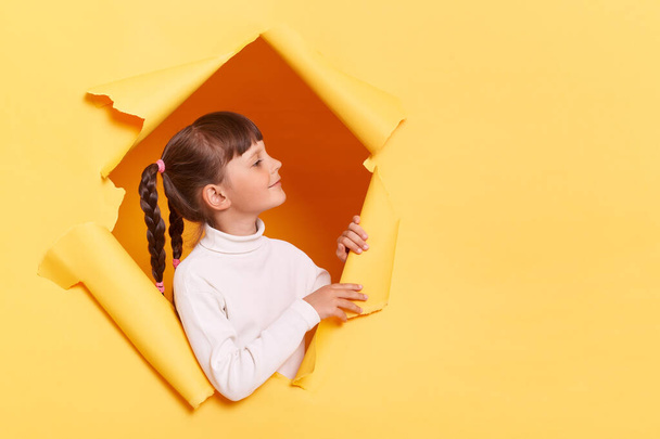 Portrait of adorable smiling little girl with braids wearing casual shirt looking through torn hole in yellow paper, looking away at copy space for promotion or advertisement. - 写真・画像
