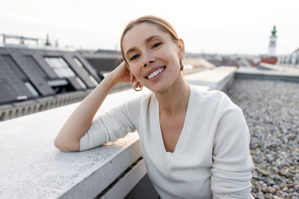 joyful woman smiling at camera while sitting on rooftop - Φωτογραφία, εικόνα