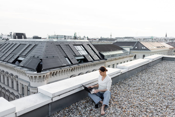 full length of barefoot woman in jeans working with laptop on rooftop of urban building - Zdjęcie, obraz