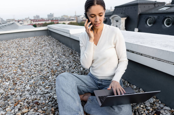 woman in white pullover sitting on rooftop, talking on smartphone and working on laptop  - Fotografie, Obrázek