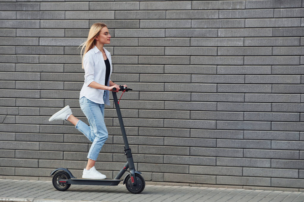 Side view of beautiful blonde in casual clothes that riding electric schooter outdoors at sunny daytime. - Photo, image