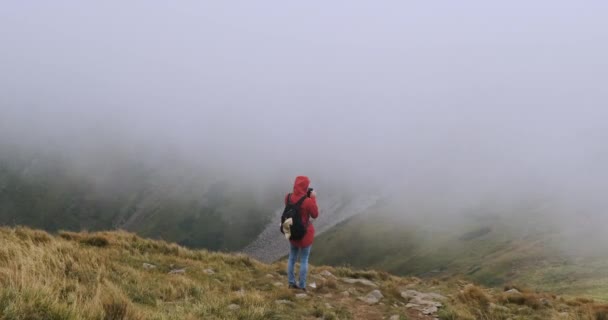 young girl with a backpack, stands in the mountains on the edge of a cliff and do something on a smartphone, online shopping, correspondence. Strong cloud cover, wind, bad weather. Carpathians - Video