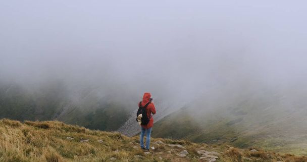 young girl with a backpack, stands in the mountains on the edge of a cliff and do something on a smartphone, online shopping, correspondence. Strong cloud cover, wind, bad weather. Carpathians - Fotó, kép