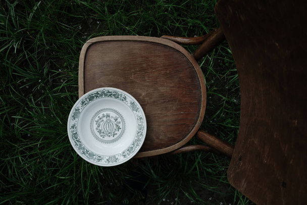 Grandma's white kitchen bowl with a green floral pattern on an old wooden chair in the garden. Cottage core concept. Dark photo. Top view. Copy space. - Photo, Image