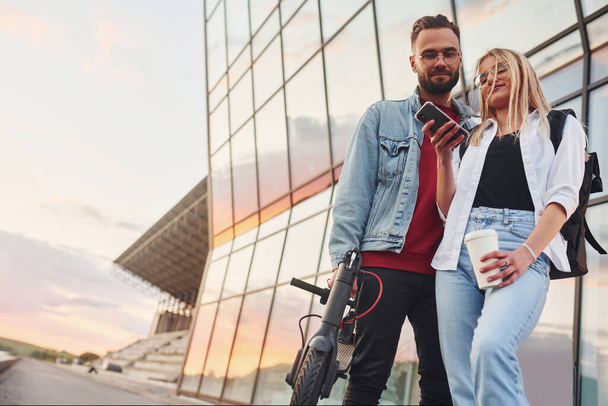 With electric schooter. Young stylish man with woman in casual clothes outdoors near business building together. Conception of friendship or relationships. - Foto, Bild