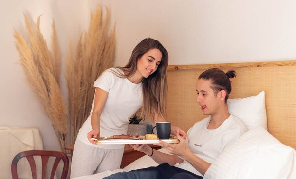 Bonito jovem casal heterossexual na cama prestes a tomar o delicioso café da manhã juntos em um quarto mínimo boho - Foto, Imagem