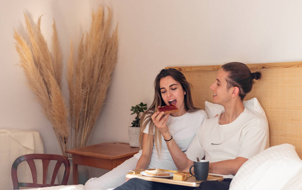 Happy young lovely man and woman couple giving food to each other while having breakfast in bed as romantic date - Fotografie, Obrázek