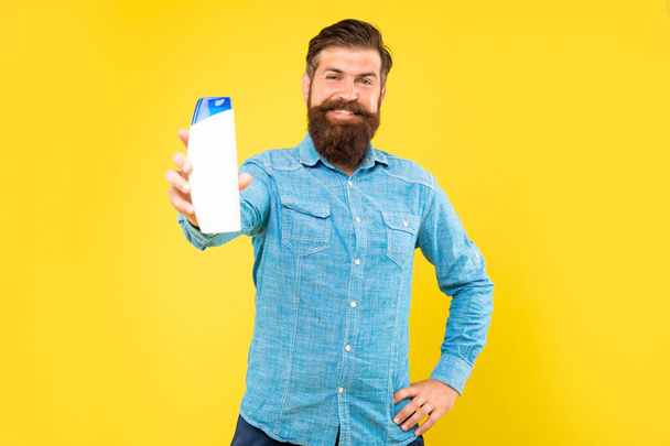 brutal bearded man hold hair conditioner bottle on yellow background, presenting toiletries - Photo, image