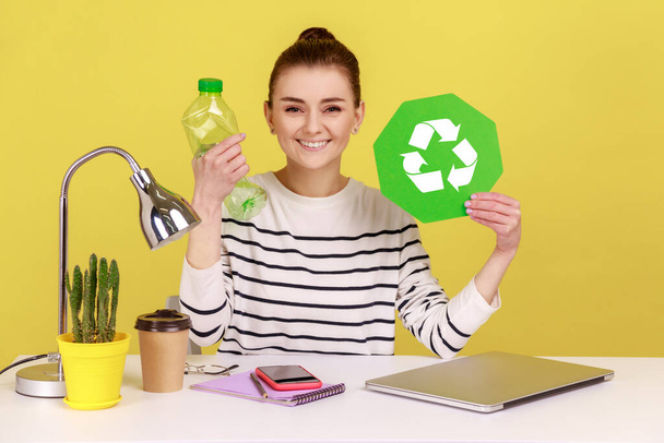 Portrait of satisfied woman with charming smile holding empty plastic bottle and green recycling sign, sitting on workplace. Indoor studio studio shot isolated on yellow background. - Foto, Bild