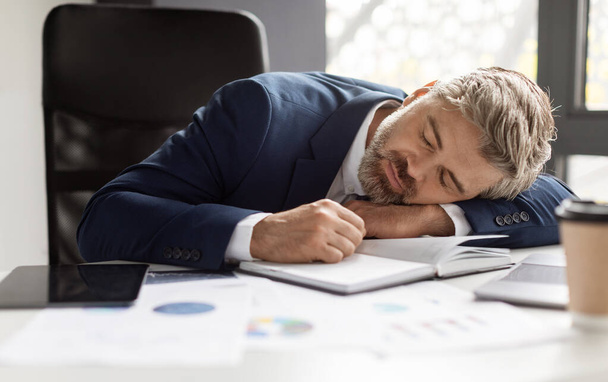 Przepracowana koncepcja. Wyczerpany dojrzały biznesmen śpiący w miejscu pracy w nowoczesnym biurze - Zdjęcie, obraz