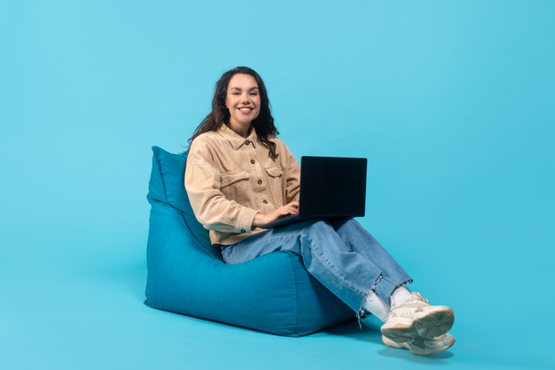 Cheerful young caucasian brunette lady sitting on bagchair and typing on laptop - Photo, Image