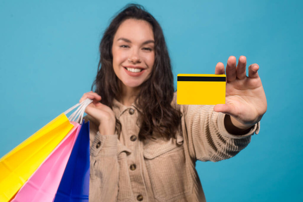 Glad young european brunette female with purchases bags showing credit card isolated on blue background - Foto, immagini