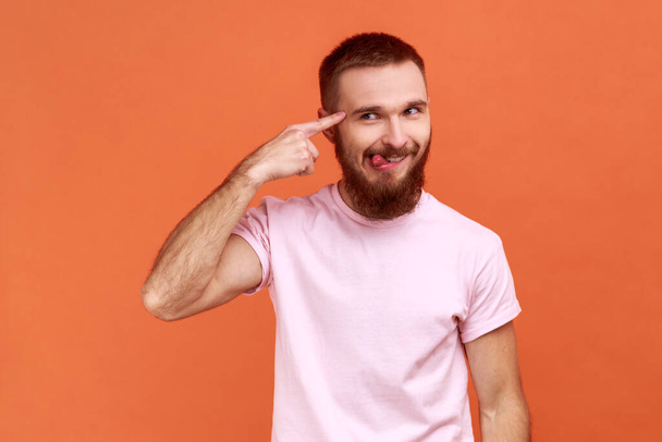 Portrait of bearded man showing stupid gesture, looking at camera with condemnation and tongue out, dumb suggestion, wearing pink T-shirt. Indoor studio shot isolated on orange background. - Foto, imagen
