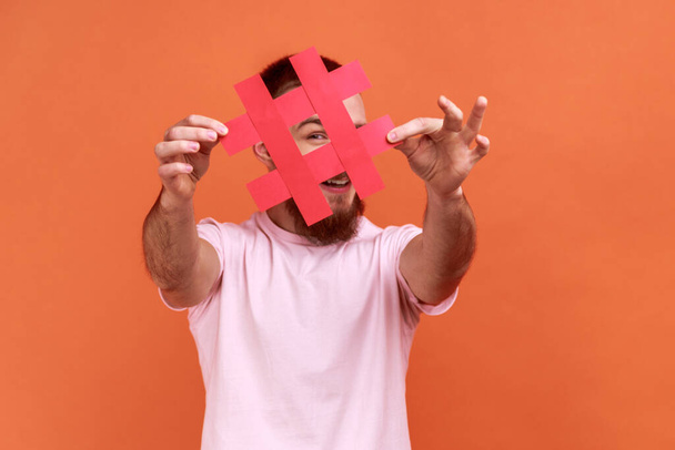 Portrait of bearded man covering face with social media hashtag symbol, recommending to follow trendy content, popular blog, wearing pink T-shirt. Indoor studio shot isolated on orange background. - Photo, image