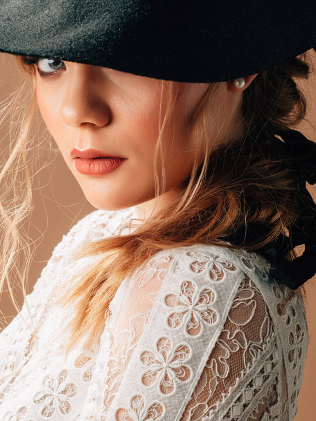 Portret van een mooie jonge blonde bruid met zwarte rustieke hoed. Cowboy trouwstijl. Elegante bruidsjurk met zacht katoenen kant en lange transparante mouwen. Studioshoot. - Foto, afbeelding