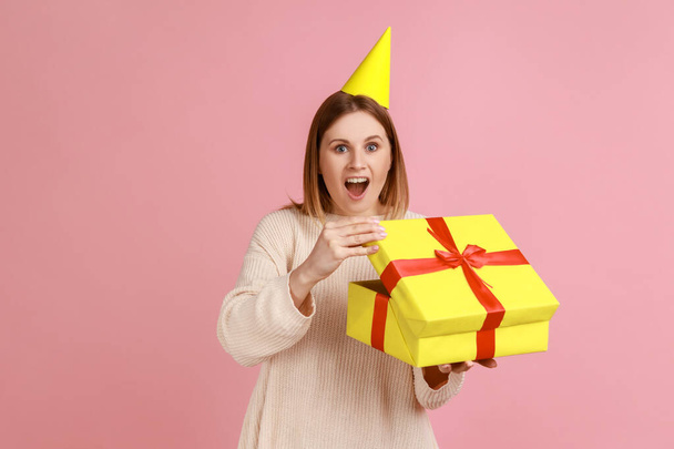 Portrait of excited astonished blond woman in party cone unpacking present box, having pleasant surprise, looking at camera, wearing white sweater. Indoor studio shot isolated on pink background. - Foto, Imagen