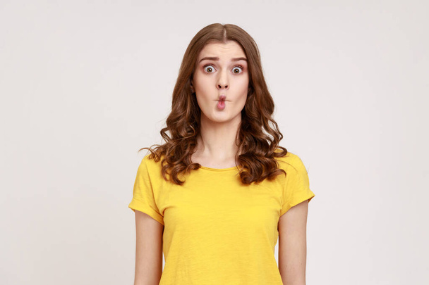 Portrait of charming funny teen girl with wavy hair in yellow T-shirt shows fish face grimace with pout lips, makes ridiculous childish comical grimace. Indoor studio shot isolated on gray background. - Photo, image