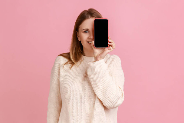Portrait of smiling blond woman covering her eye with smartphone with empty display for advertisement, having playful expression, wearing white sweater. Indoor studio shot isolated on pink background. - Photo, image