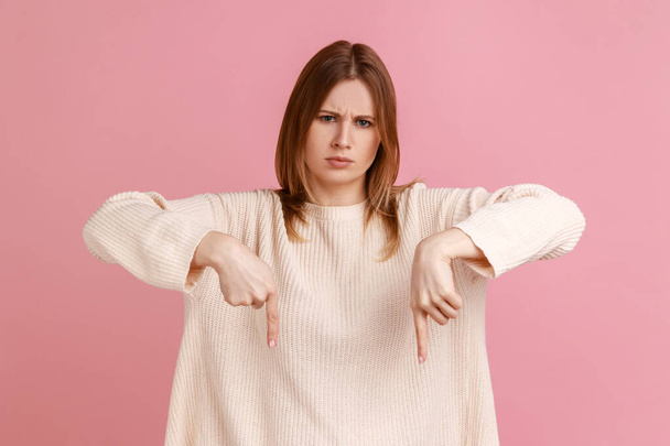 Portrait of strict bossy blond woman showing place down beside her, giving order with strict bossy expression, wearing white sweater. Indoor studio shot isolated on pink background. - Fotó, kép