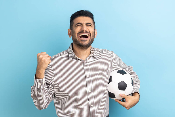 Excited businessman screaming widely opening mouth, celebrating victory of favourite football team, holding soccer ball in hands, wearing striped shirt. Indoor studio shot isolated on blue background. - Fotoğraf, Görsel
