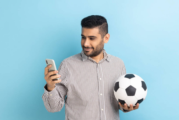 Portrait of smiling optimistic handsome bearded businessman standing with soccer ball and using mobile phone, wearing striped shirt. Indoor studio shot isolated on blue background. - Foto, afbeelding